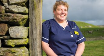 a nurse leaning on a wooden gatepost next to a drystone wall with green countryside behind her