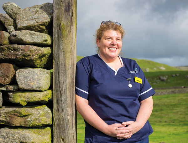 a nurse leaning on a wooden gatepost next to a drystone wall with green countryside behind her