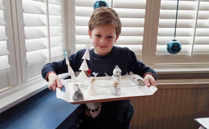 A small boy holds a tray with homemade figures and trees
