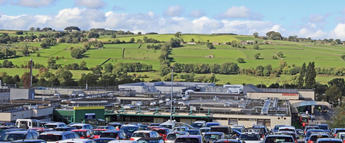 a hospital and car park with rolling countryside with trees and drystone walls in the background