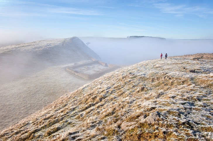 Two walkers at the top of a hill shrouded in mist with Hadrian's Wall and a milecastle visible below