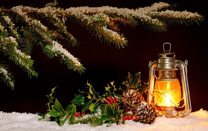 Christmas scene - an oil filled lantern burning bright with snow covered tree, holly and ivy lit up by the glow of the lamp.