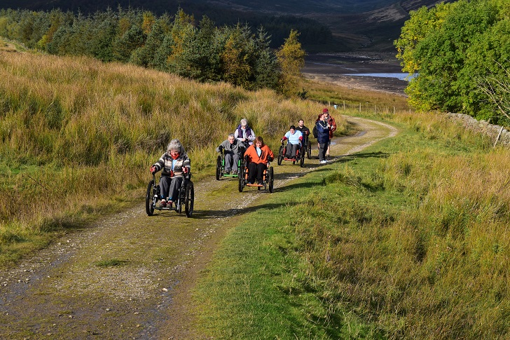 a group of wheelchair users heading up an off-road path in upland countryside near a reservoir