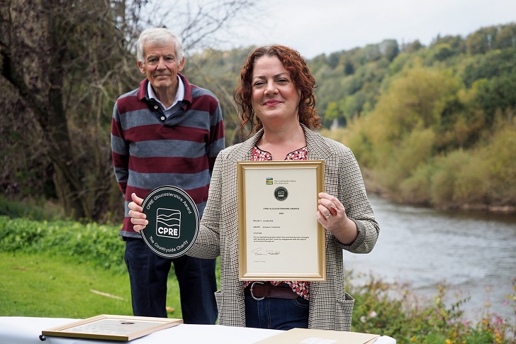 A man standing behind a woman holding an award plaque and certificate in a rural riverside setting
