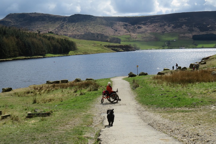 a woman in a wheelchair with a dog on a path leading to a reservoir surrounded by green hills