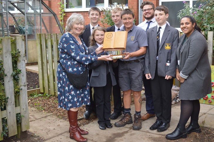 A woman presenting a carved wood award to a group of school children in a garden area