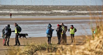 walkers on a beach with sand dunes i nthe foreground and wind turbines beyond