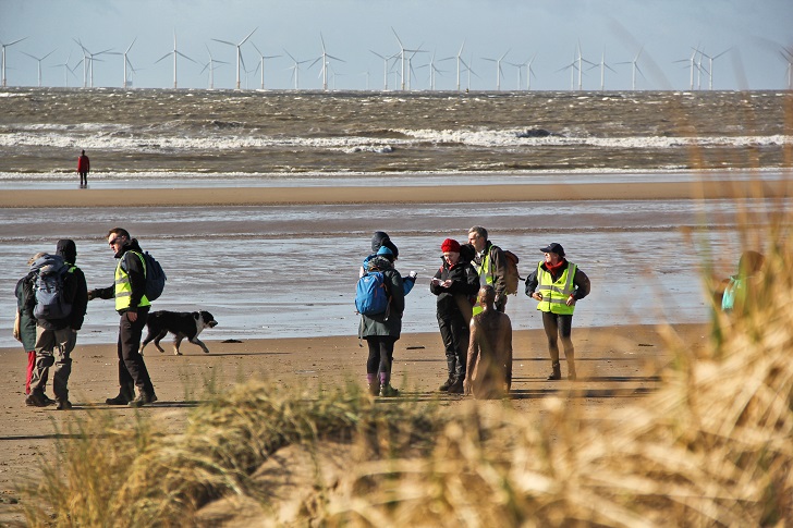 walkers on a beach with sand dunes i nthe foreground and wind turbines beyond