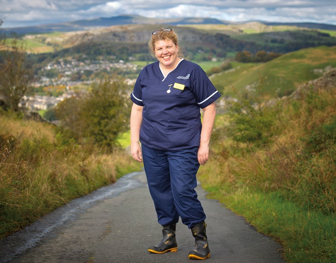 A nurse in uniform standing in a steep country lane with a town in a rural valley behind her