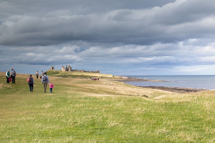 Families walking along a grassy coastline with the sea and a beach on the right and a ruined castle in the distance