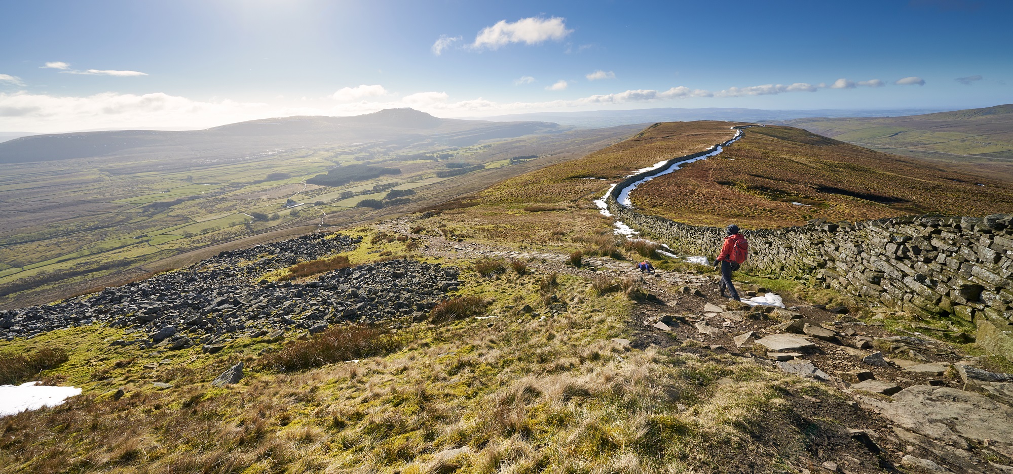 A walker descending a hillside path in bright sunshine with snow and scree on the ground