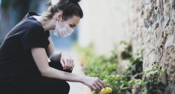 A woman wearing a mask squats down to touch a flower in a flowerbed
