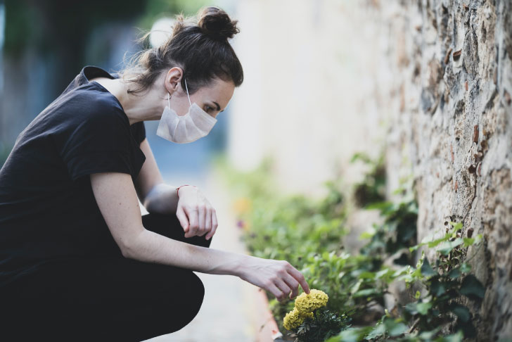A woman wearing a mask squats down to touch a flower in a flowerbed