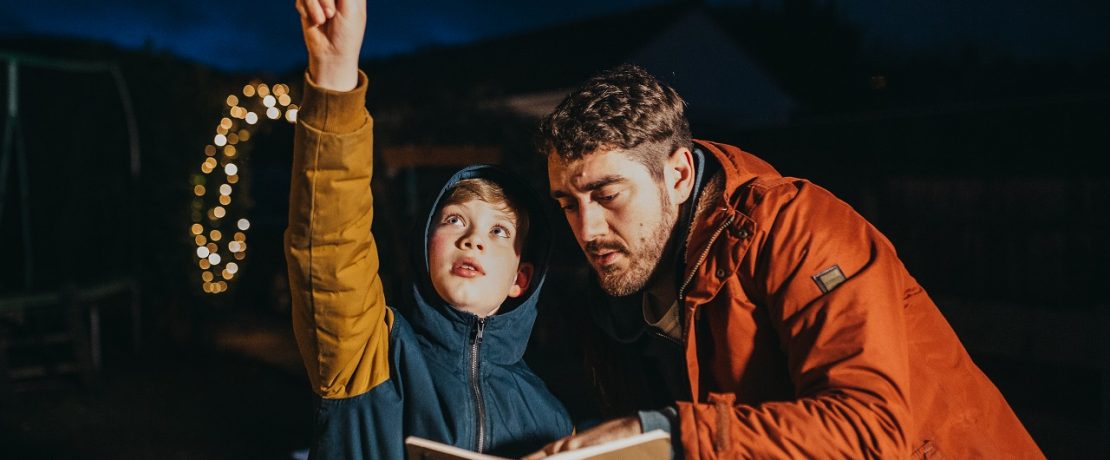 A young boy points to the night sky as his dad looks at a notepad beside him