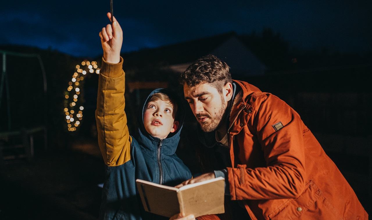 A young boy points to the night sky as his dad looks at a notepad beside him
