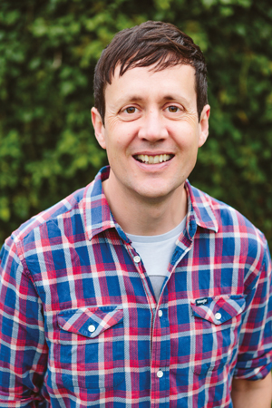 A man in a checked shirt smiling in front of foliage