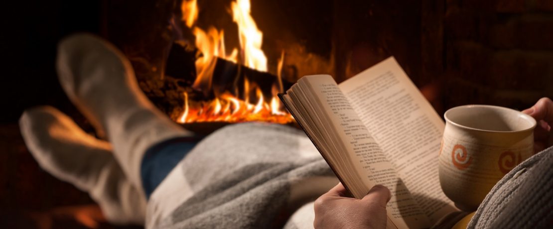 a woman reading a book in front of a log fire holding a hot drink