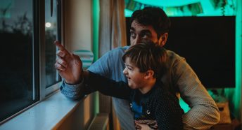 A father and son look out of a window from inside a house to a dark sky