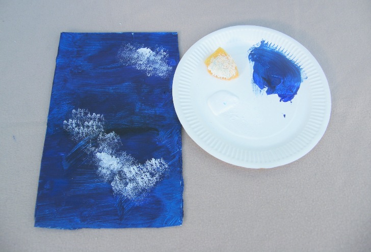 A sponge with white paint being used to make clouds onto blue card
