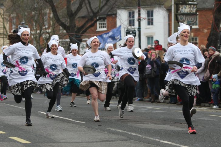 Women in aprons and scarves running with frying pans down high street in Olney