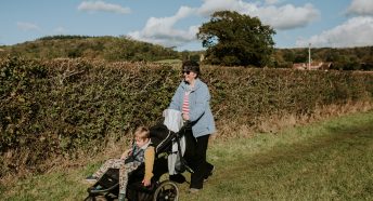 Woman pushing pushchair with small child in it alongside hedge in field