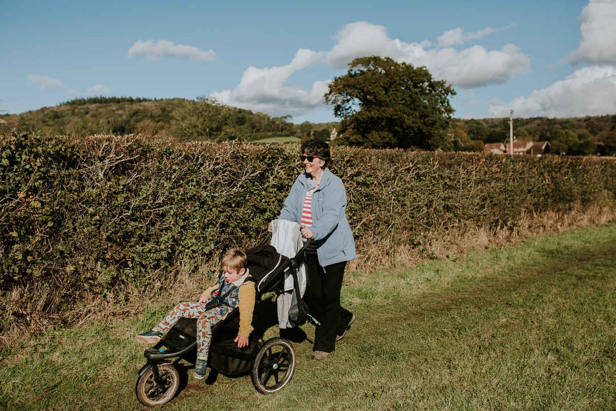 Woman pushing pushchair with small child in it alongside hedge in field