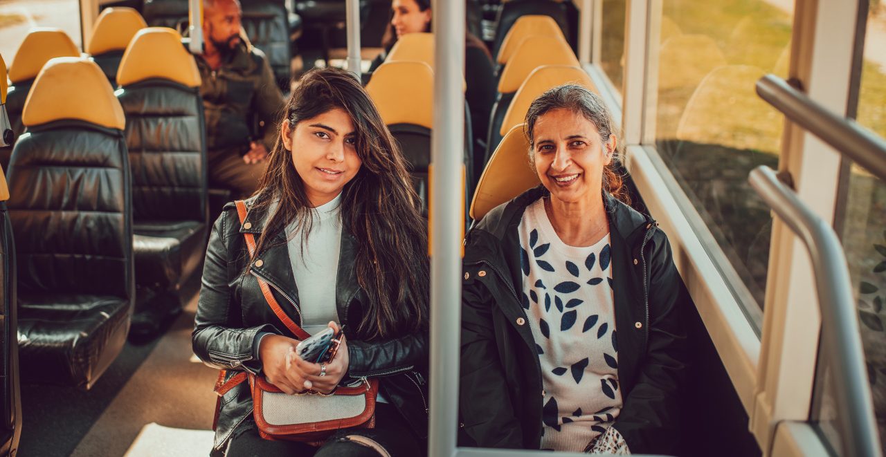 Two women sit on a rural bus service and face the camera