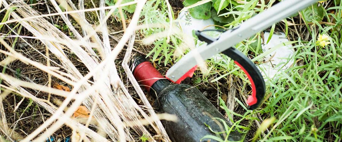 A discarded beer bottle in grass being picked up by a litter pincer