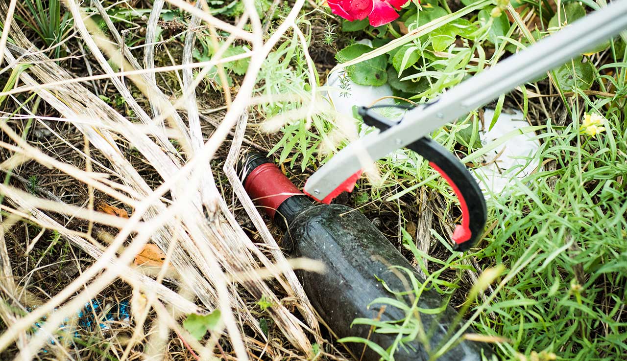 A discarded beer bottle in grass being picked up by a litter pincer