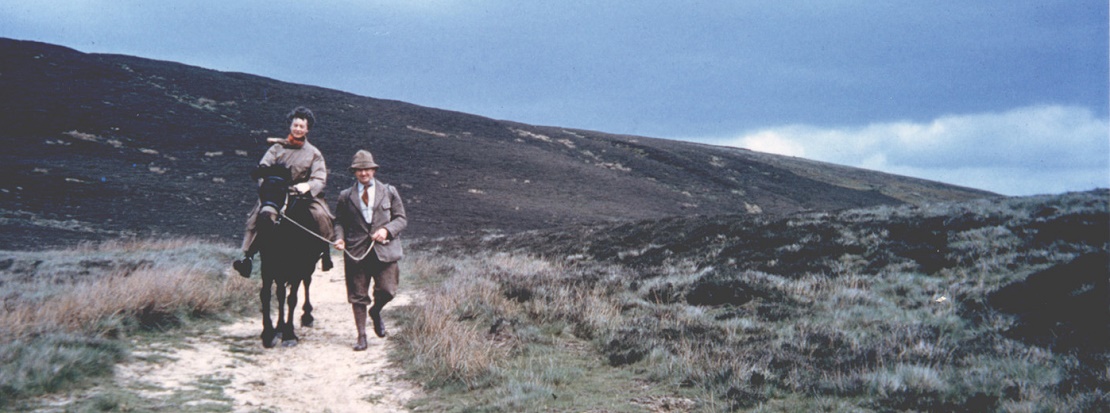 A woman on a pony led by a man on an upland hillside