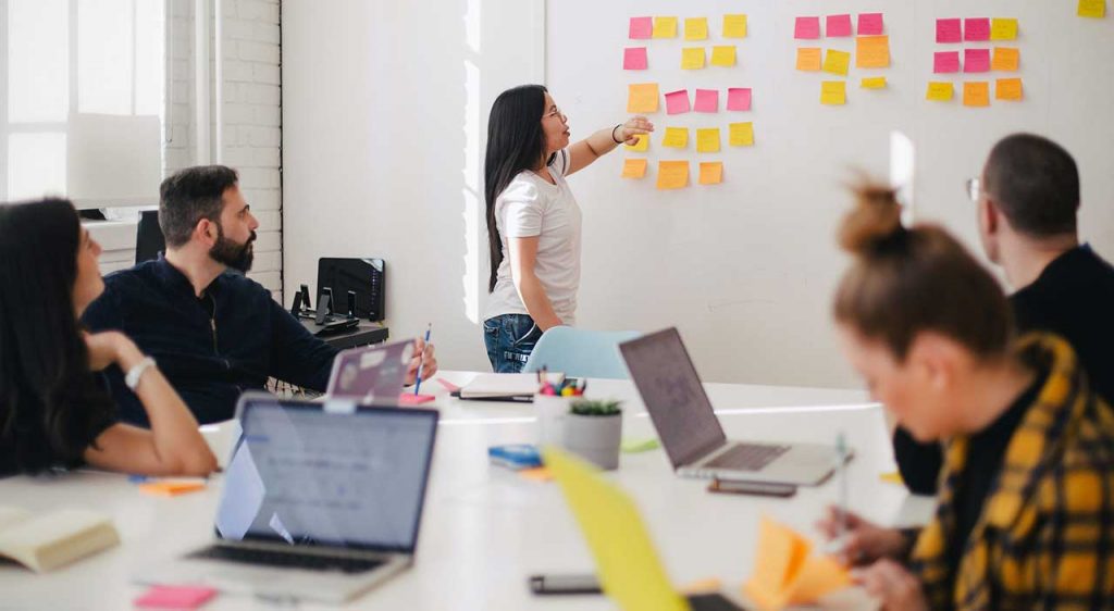 A woman stands at a white wall with post-its across it while others look on