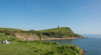 A hilly green coastline with the sea and a tower in the background