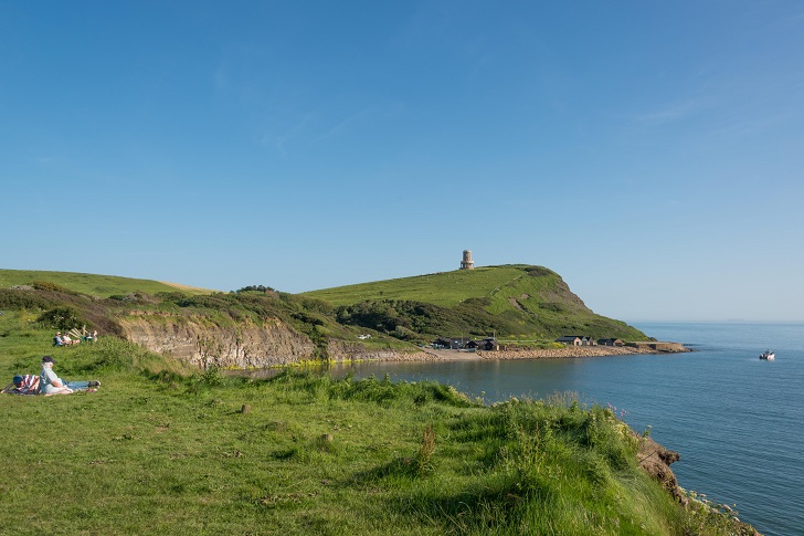 A hilly green coastline with the sea and a tower in the background