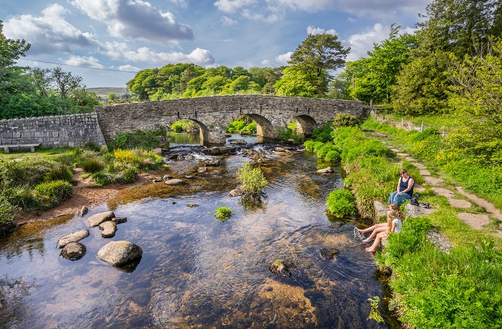 A mother and daughters sitting by a gentle river with a stone bridge in background