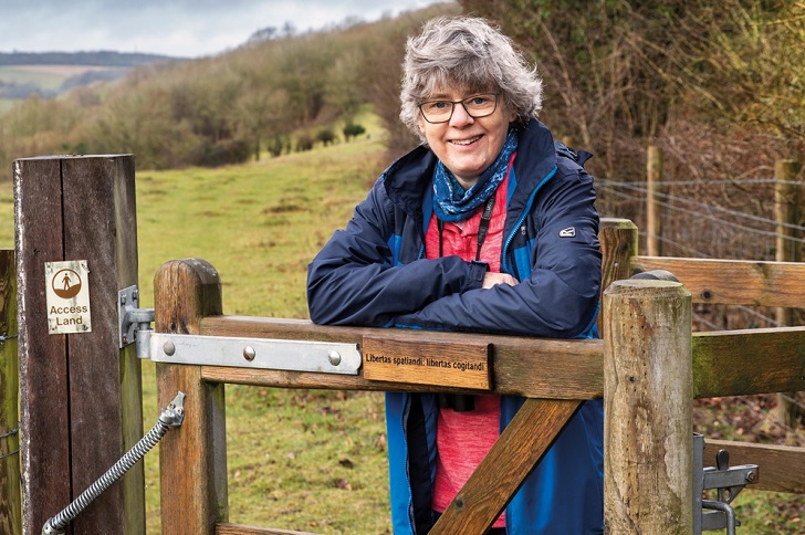 A woman in an anorak leaning on a gate with green hills in the background