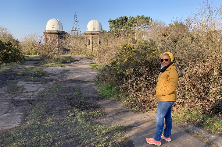 A woman standing in front of an observatory building shrouded in trees and gorse