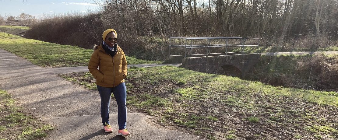 A woman on a footpath by a stream with a small footbridge in the background