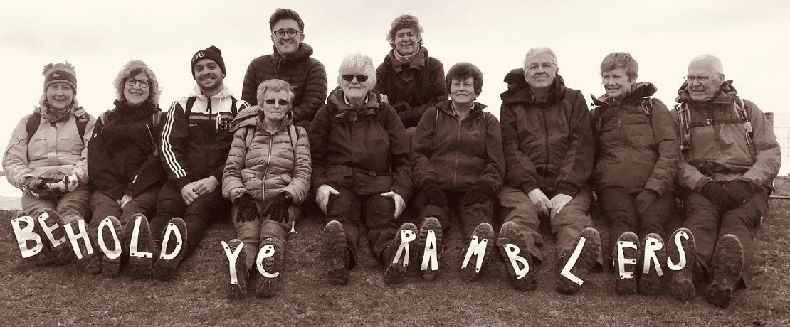 Black and white photo of row of walkers sitting on top of hill ridge with the works Behold Ye Ramblers attached to the soles of their boots