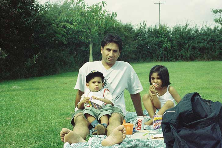 A young South Asian man sits with a small boy on his lap and little girl beside eating a picnic