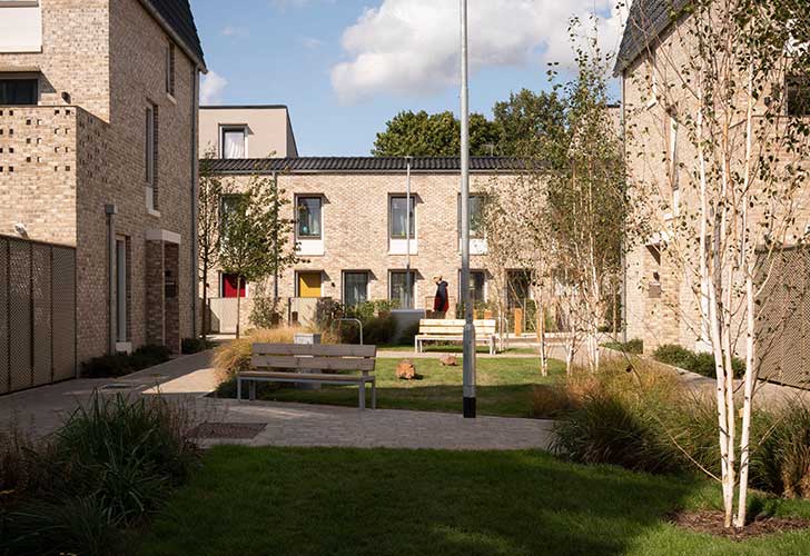 A bright sunny courtyard with benches and grass with pale stone houses around