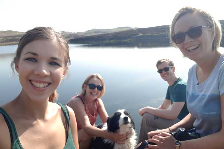 Three women and a young man sit with a border collie dog in front of a small lake