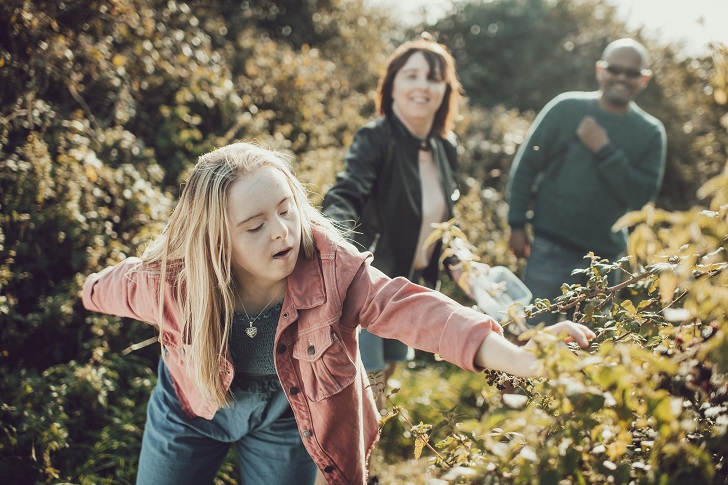 A girl picking blackberries from a hedgerow watched by her parents