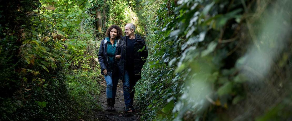 A man and a woman walk arm in arm down an overgrown path