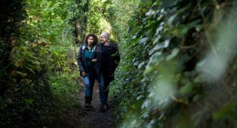 A man and a woman walk arm in arm down an overgrown path