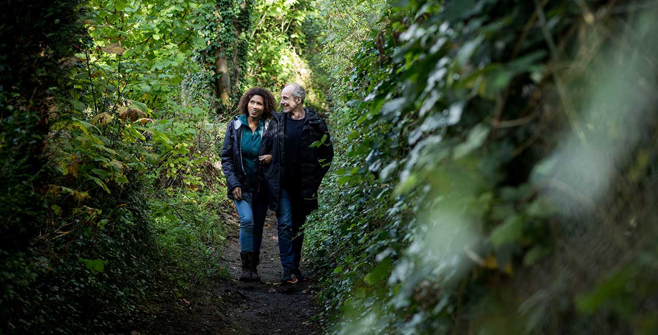 A man and a woman walk arm in arm down an overgrown path