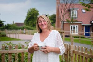 A woman holds a cup of tea and stands proudly in front of a rural home