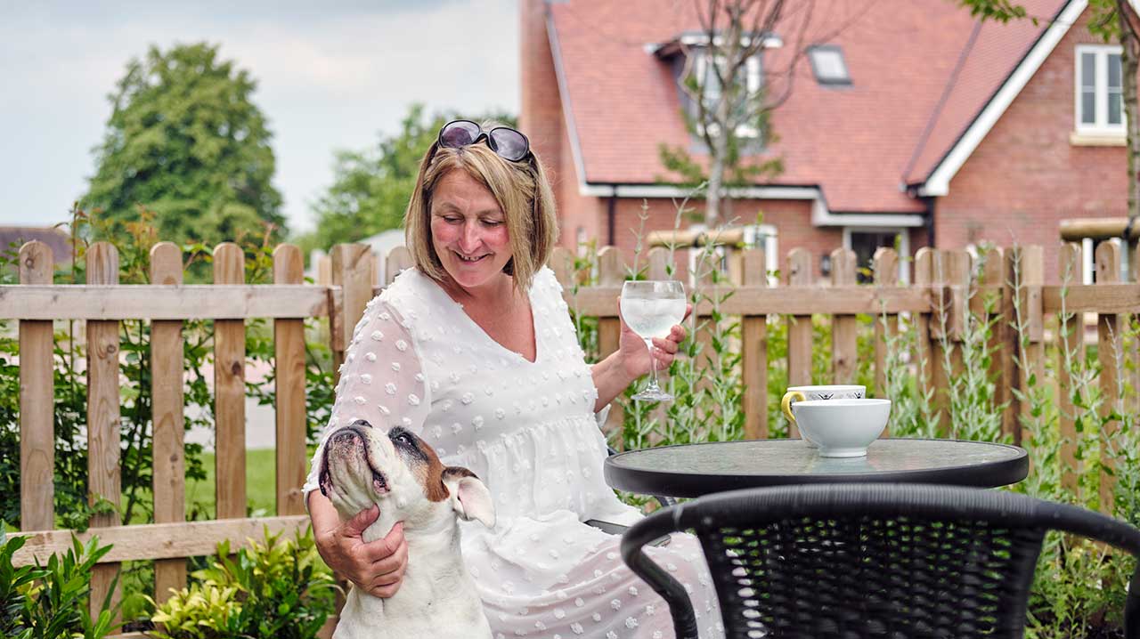 A woman sits in a garden with houses visible behind and pets a dog