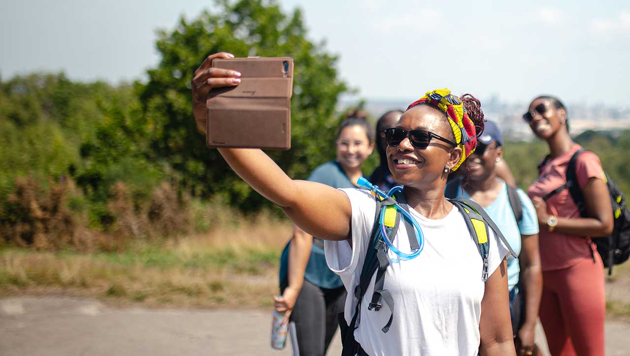 A black woman holds up her phone for a selfie with other women behind and a countryside view