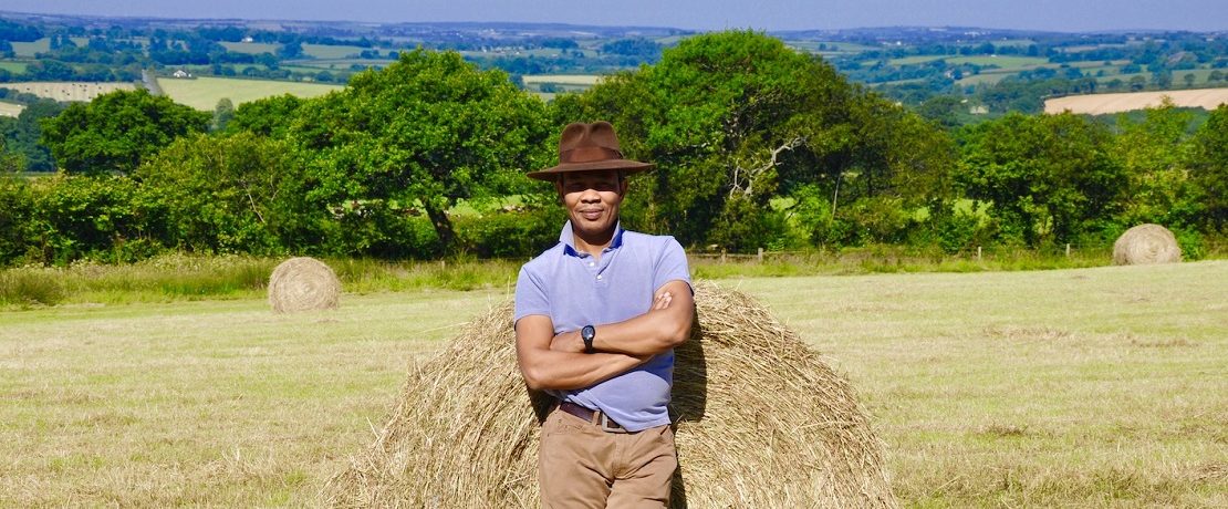 A black farmer standing in front of a round hay bale in rolling fields, hedgerows and trees