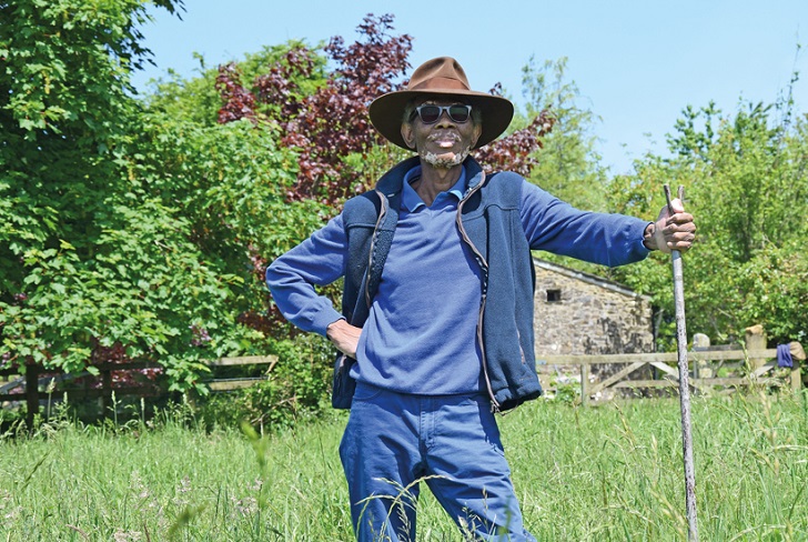 A black man leaning on a wooden staff in a grass meadow